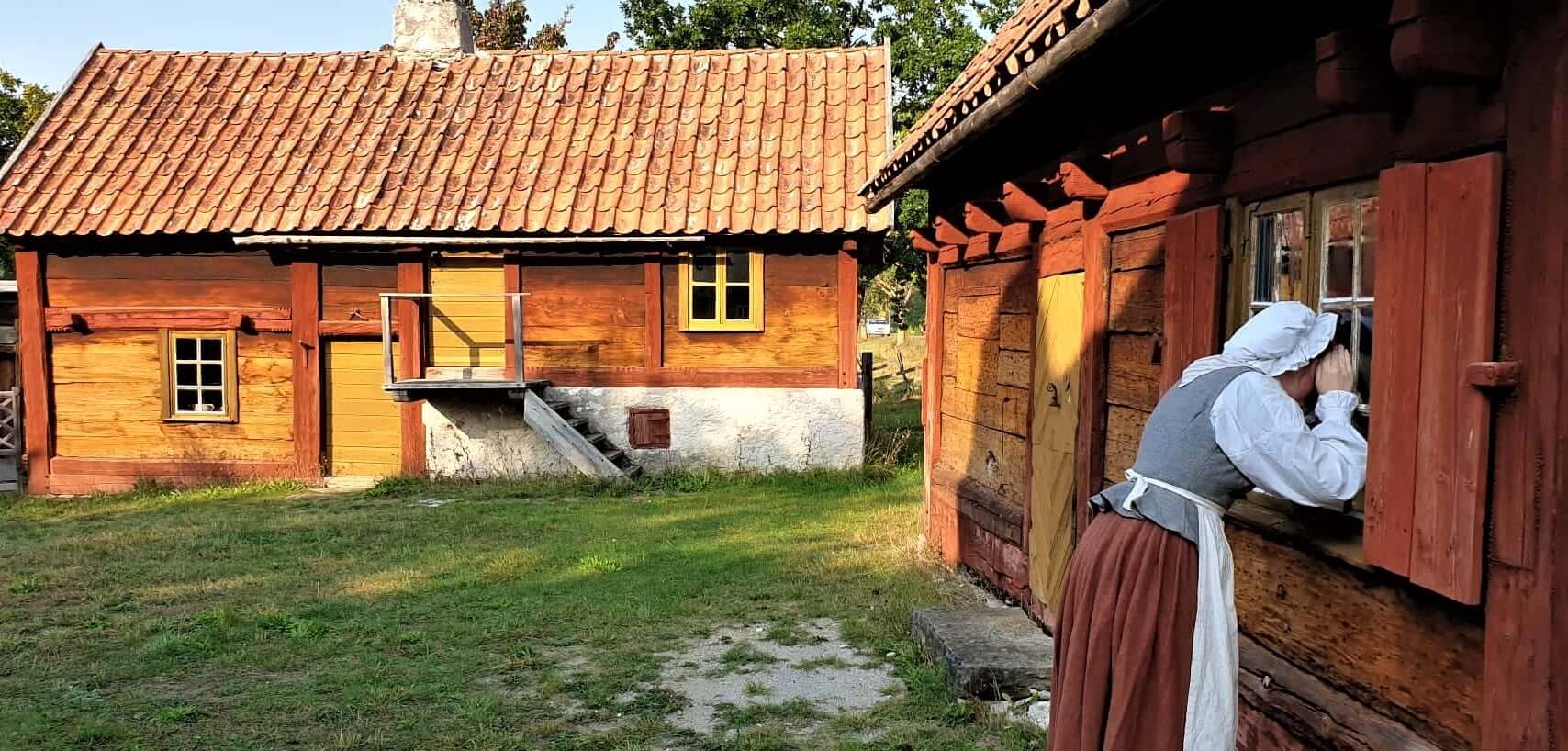 En person i historiska kläder kikar genom fönstret på ett trähus, med ett annat rustikt hus och grönt gräs synligt i bakgrunden, vilket antyder en historisk eller lantlig bymiljö.