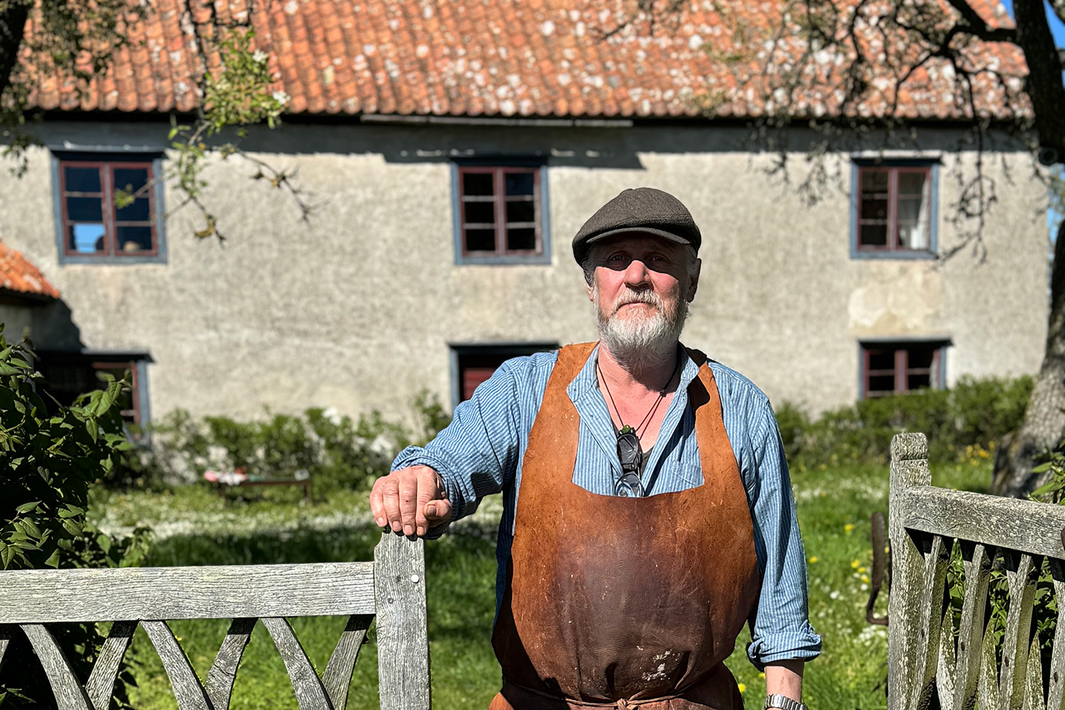 En skäggig man i platt keps, blå skjorta och brunt läderförkläde står utomhus lutad mot ett trästaket, med ett rustikt hus och grönska i bakgrunden.
