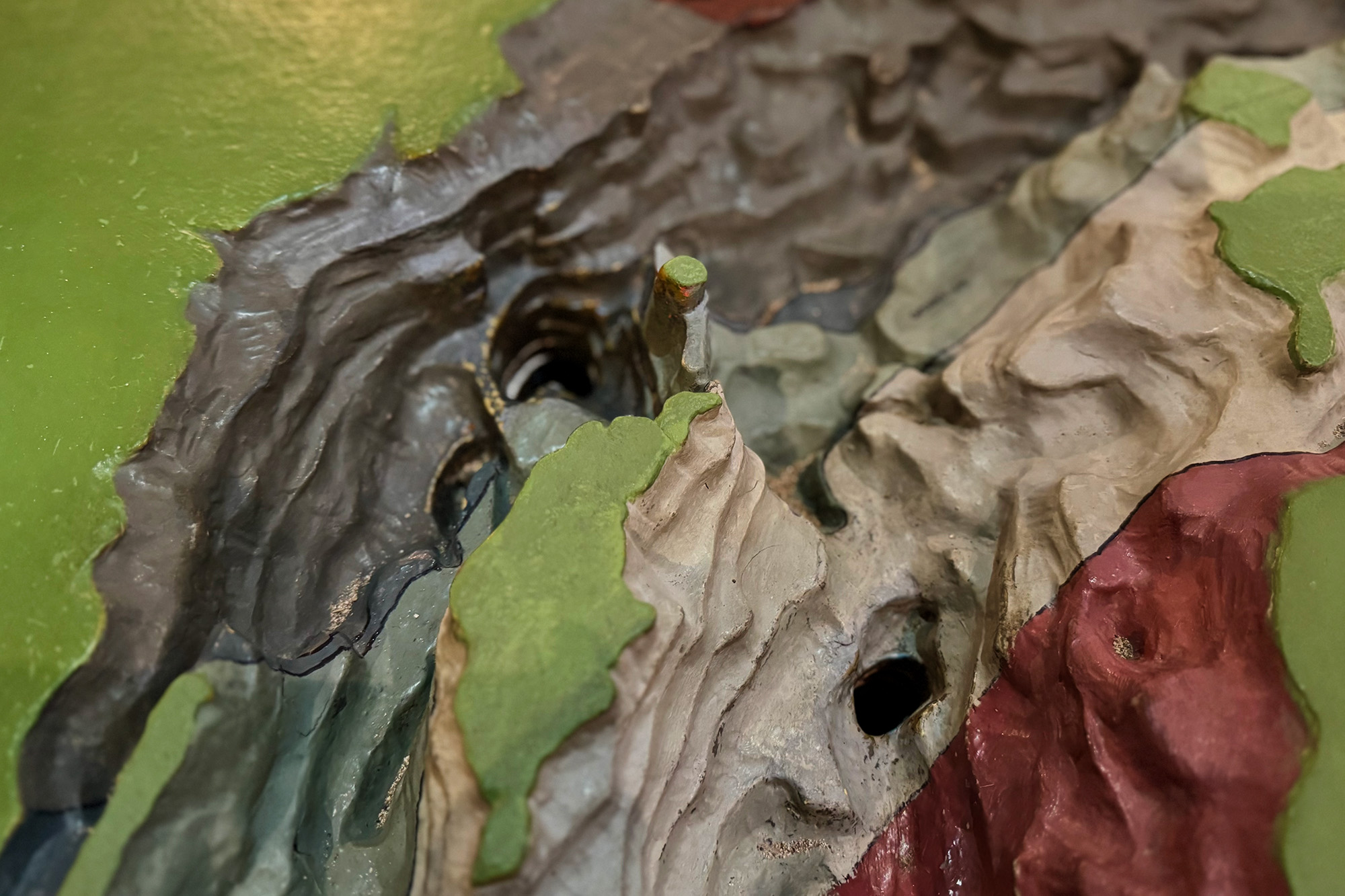 En närbild av en färgglad topografisk modell som visar dalar, klippor och platåer med gröna, grå och röda områden, och två synliga hål som representerar geologiska drag eller grottor.