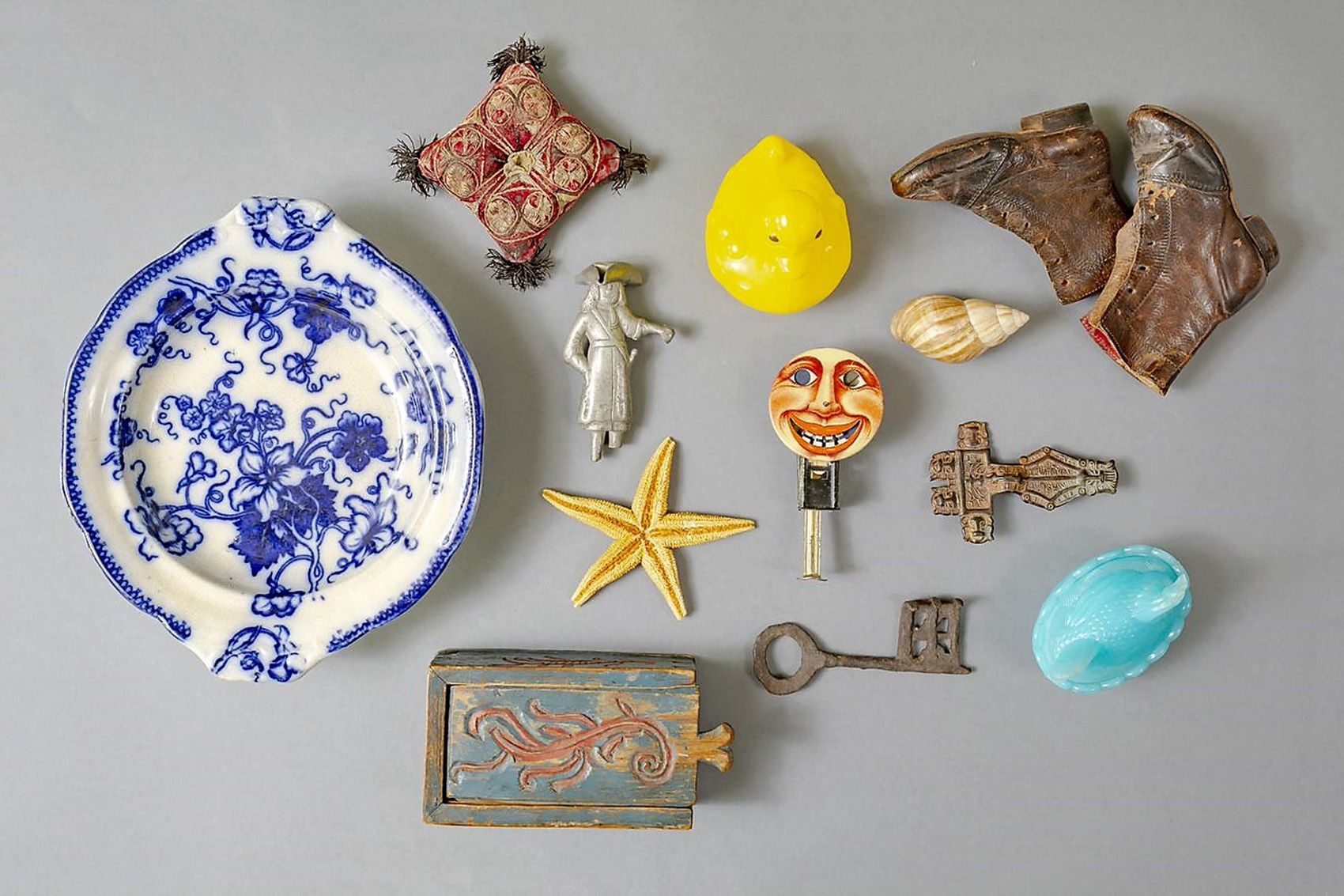 En samling vintageföremål mot grå bakgrund, inklusive en blå och vit tallrik, liten kudde, gul fågel, bruna stövlar, figurin, snäckor, nyckel med månansikte, sjöstjärna, metallföremål och en blå oval prydnadsdoska.