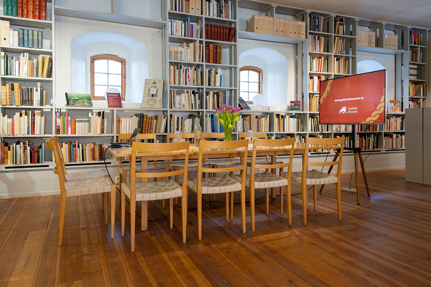 Ett väl upplyst biblioteksrum med trägolv, ett långt bord och åtta stolar, bokhyllor fyllda med böcker, två välvda fönster, en vas med blommor på bordet och en skärm som visade en webbplats.