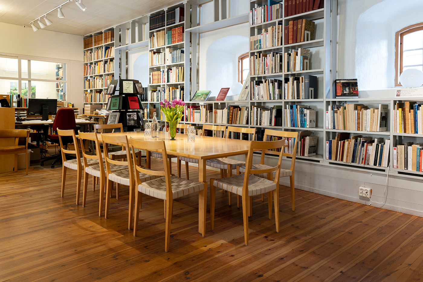 Ett mysigt biblioteksrum med höga bokhyllor fyllda med böcker, ett träbord omgivet av åtta stolar, en vas med tulpaner på bordet och trägolv. Skrivbord och kontorsutrymme syns i bakgrunden.