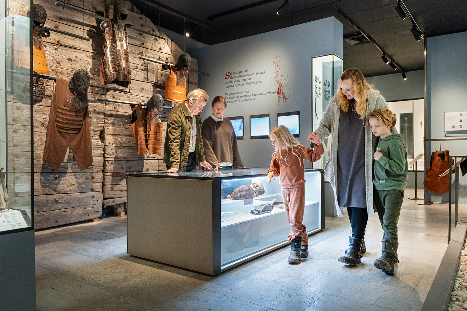 En familj med två barn och två vuxna tittar på historiska artefakter som visas i en glasmonter på en museiutställning, omgiven av trä- och tygföremål monterade på väggarna.